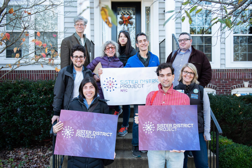 Group photo of nine NYC volunteers holding Sister District signs while standing on a porch and canvassing voters